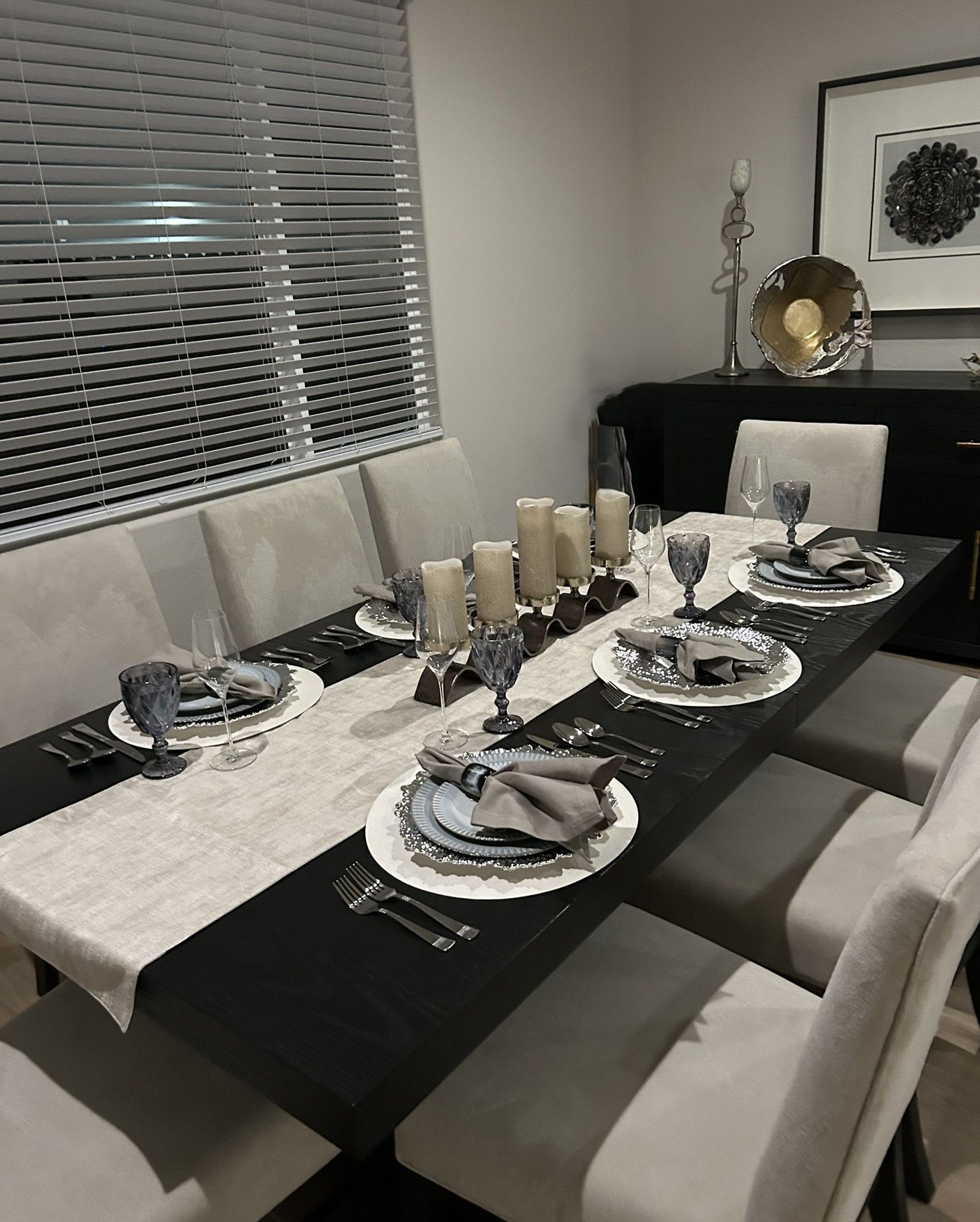 A modern dining room with a long black table set for a formal dinner, featuring cream-colored chairs and a light runner.