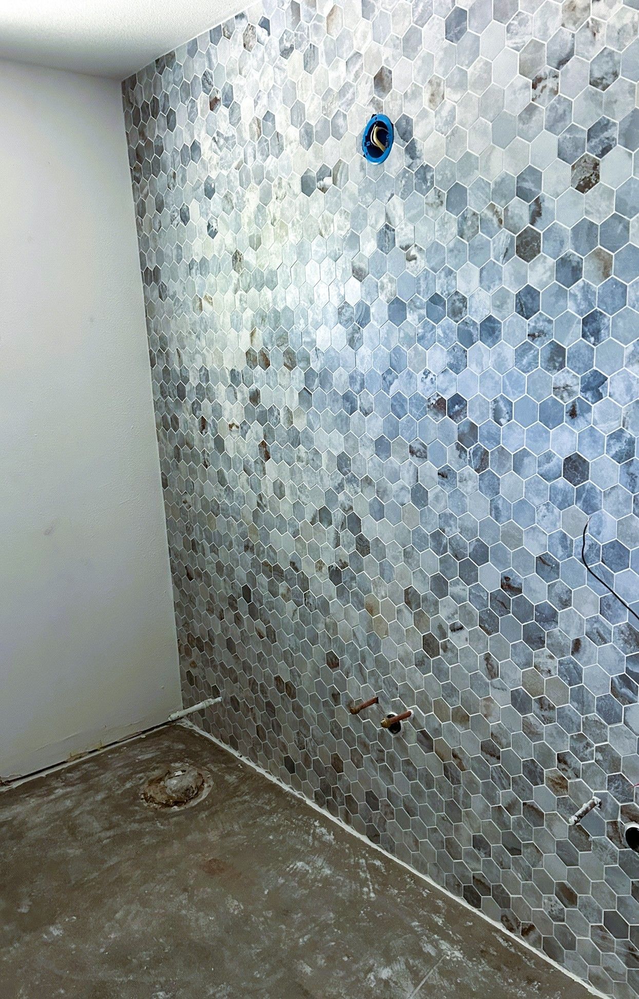 A wall tiled with gray and white marble hexagons under construction, with exposed pipes and a bare concrete floor.