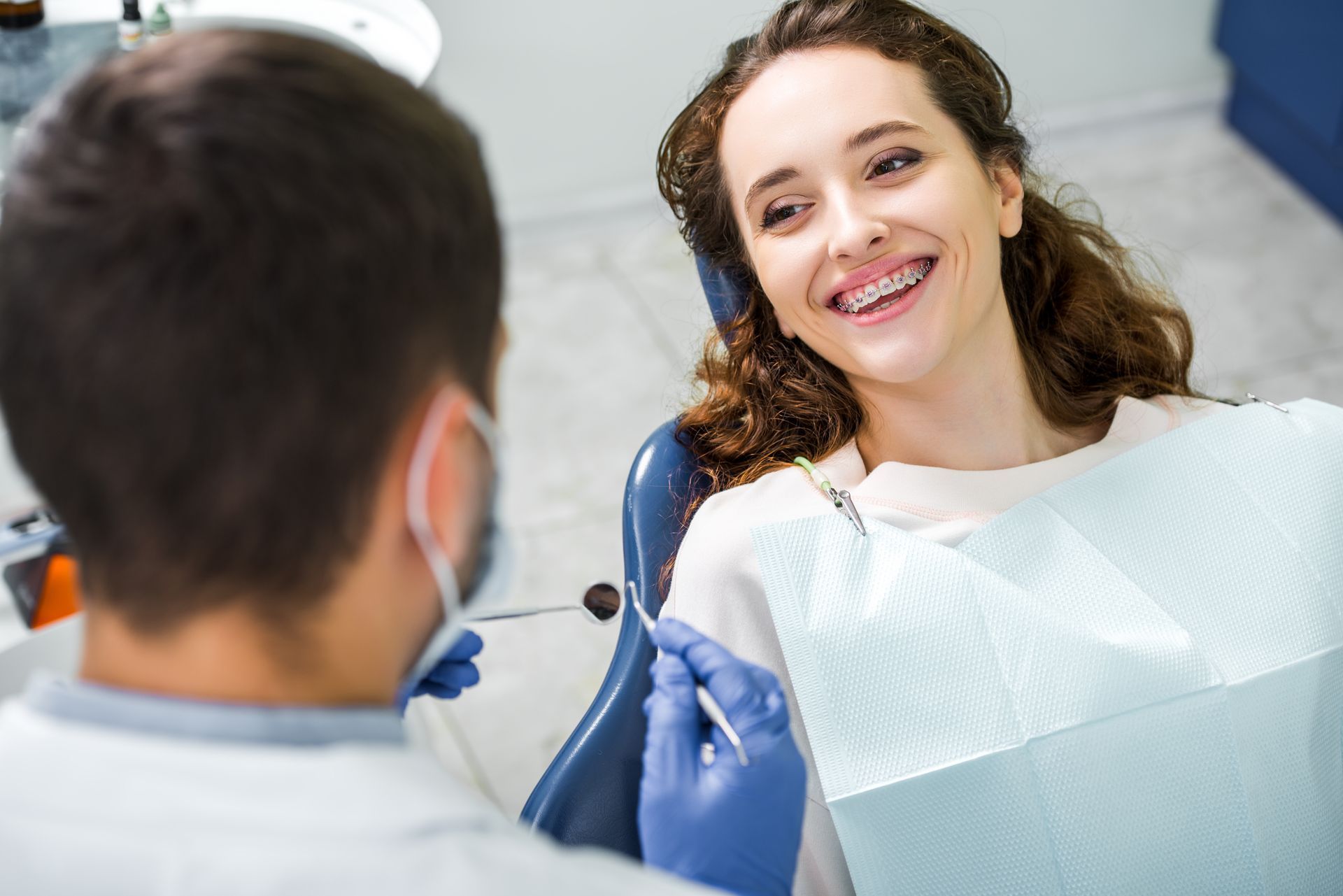 A dentist examining a patient's teeth with tools in a dental office. The patient smiles, wearing braces.
