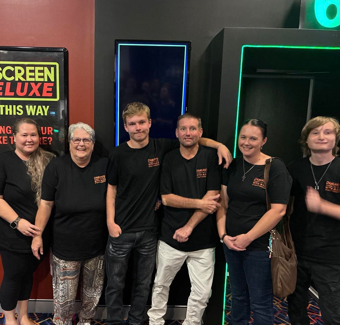 Group of six people in black shirts posing together inside a neon-lit arcade or cinema lobby