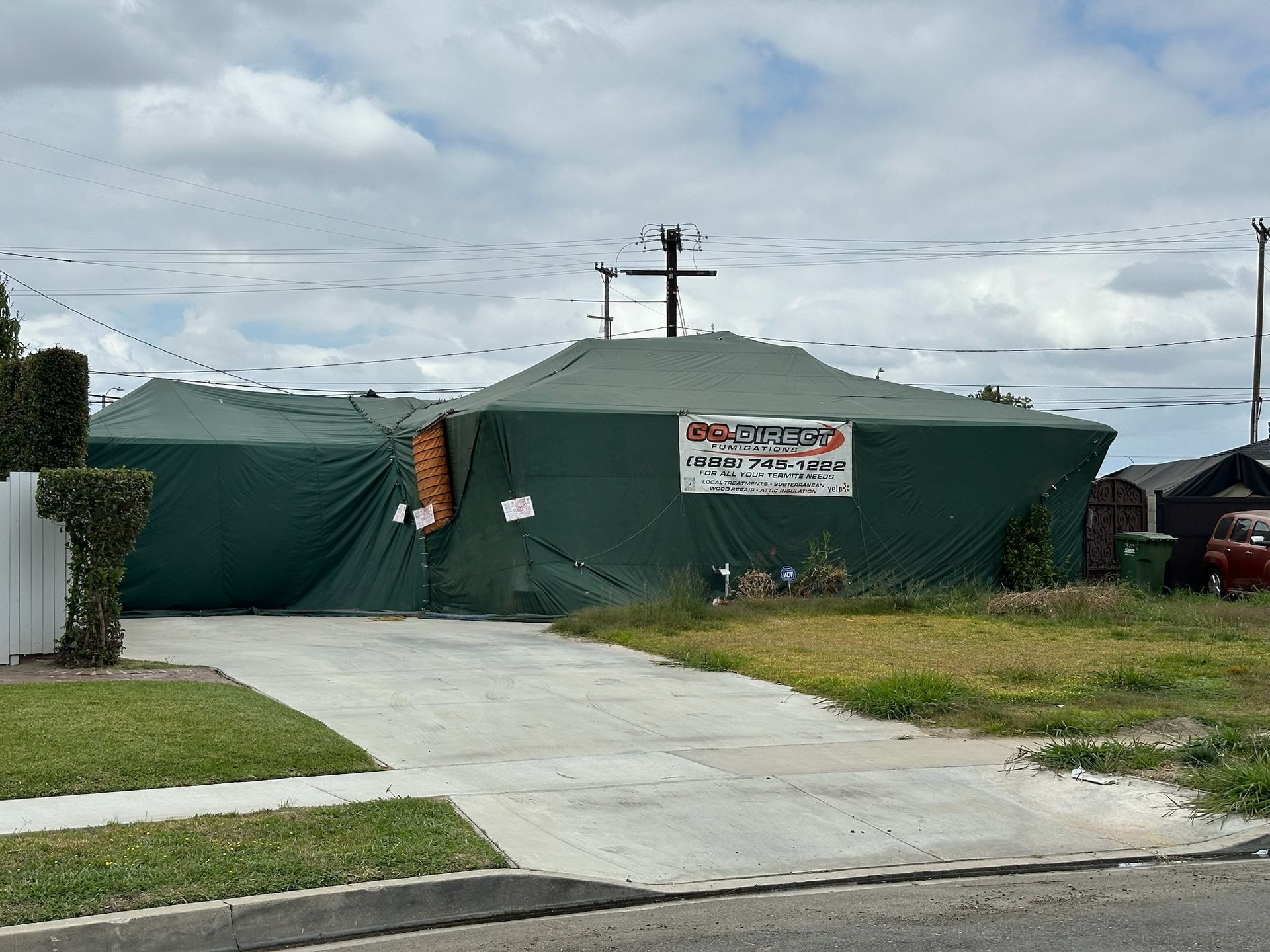 House covered in green tarpaulin for pest control, driveway in front.