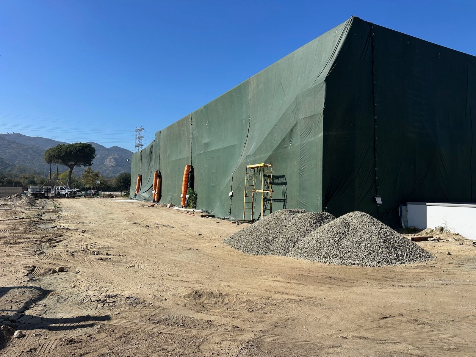 Large green tarp-covered structure in a dirt lot with piles of gravel, under a blue sky.