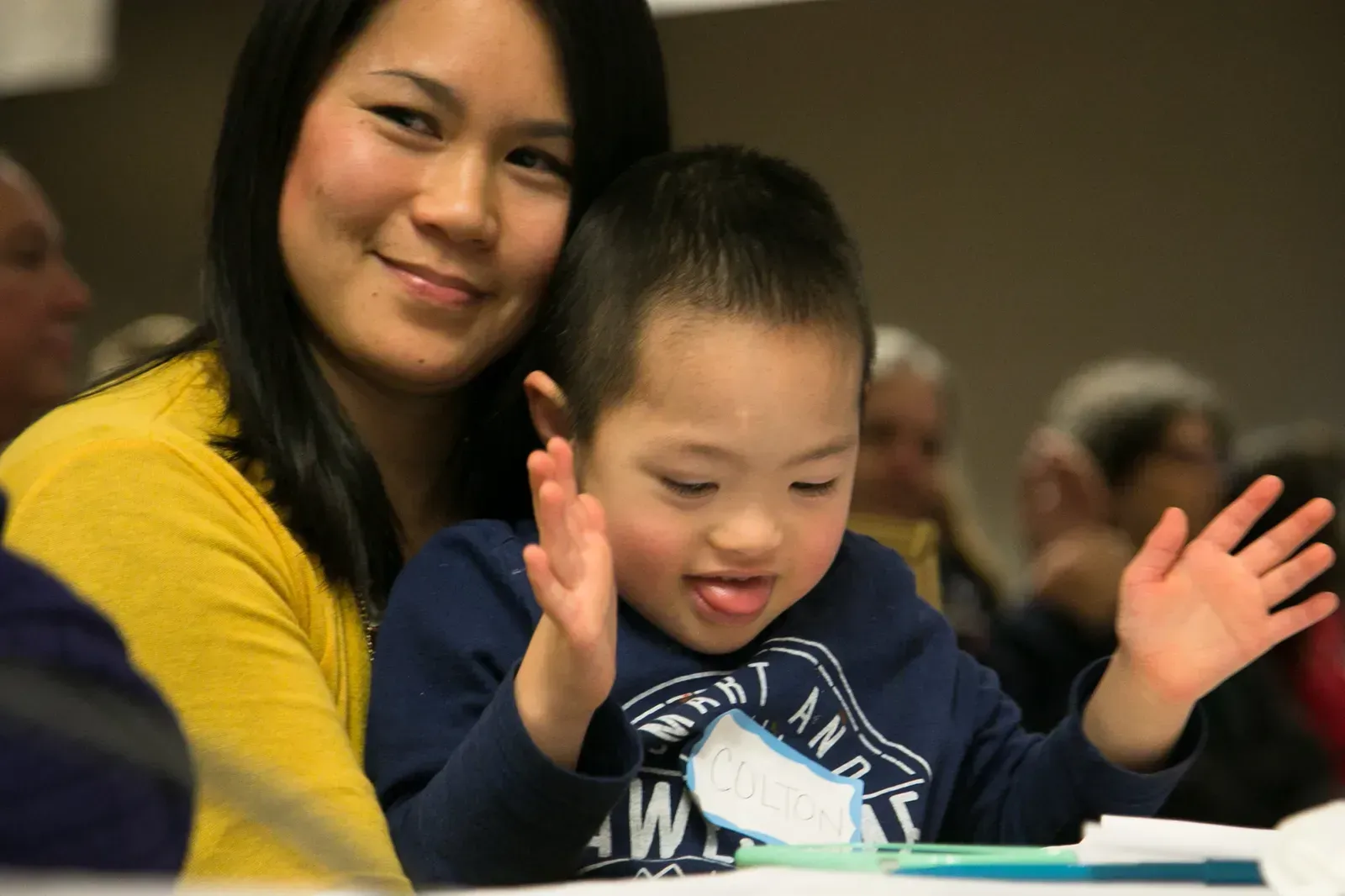 Woman smiles, holding a child with open hands; he has his tongue out. Indoor setting with others present.