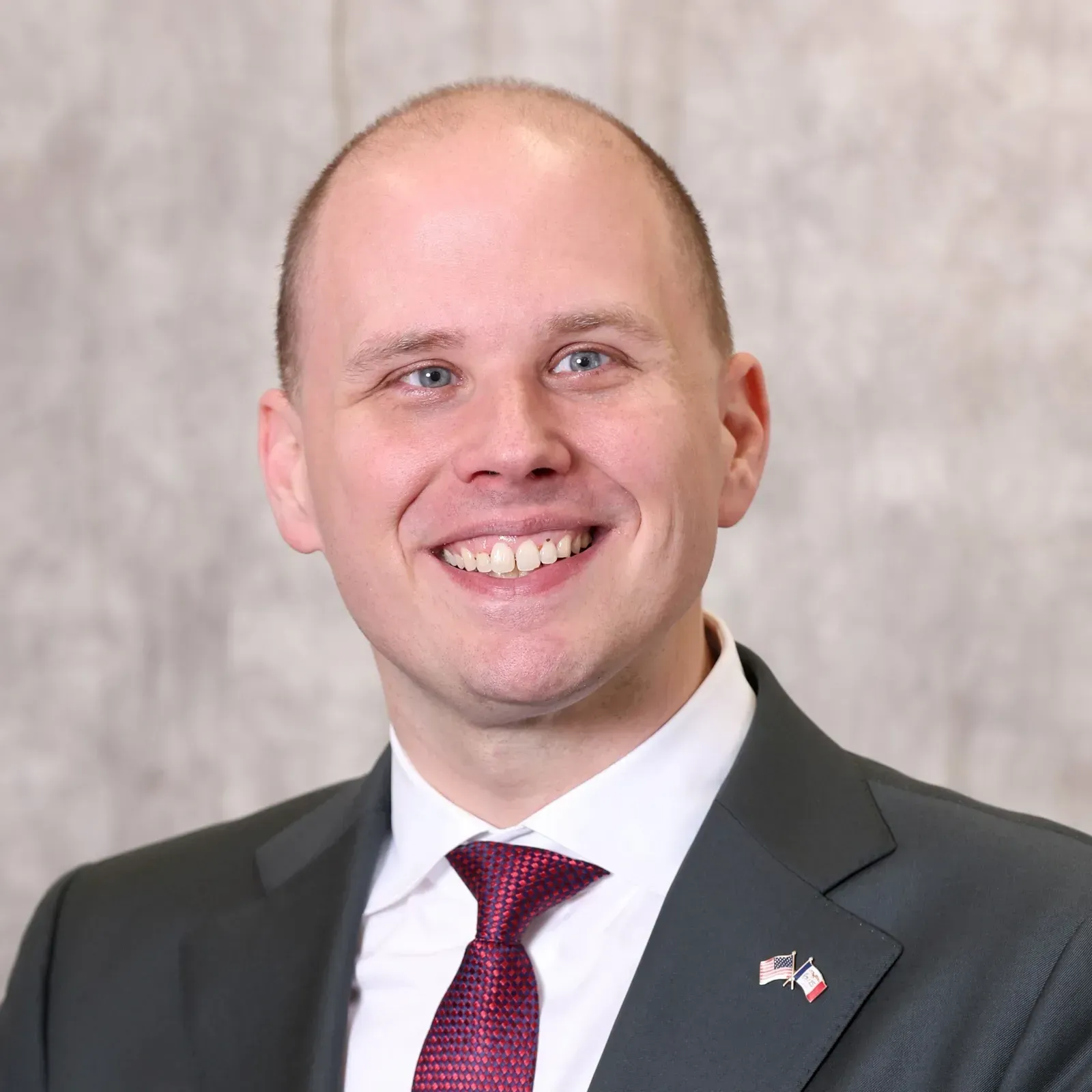 Smiling man in dark suit and maroon tie; backdrop with texture.