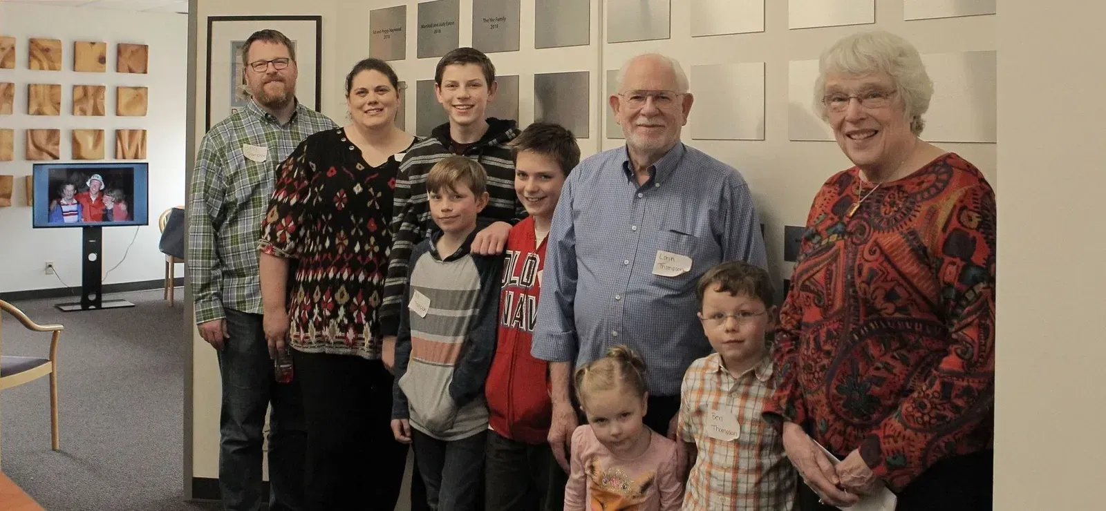 A group of people, including children and seniors, pose for a photo in front of a wall of plaques.