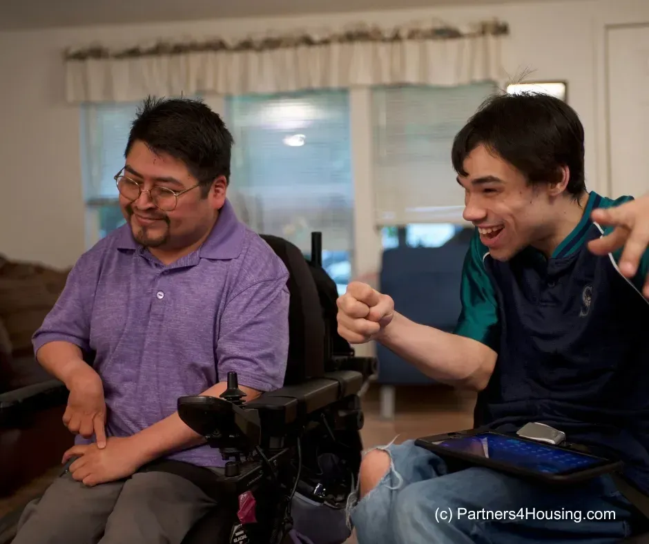 Two men in wheelchairs, one smiling, the other pointing excitedly. Indoors, near a window.