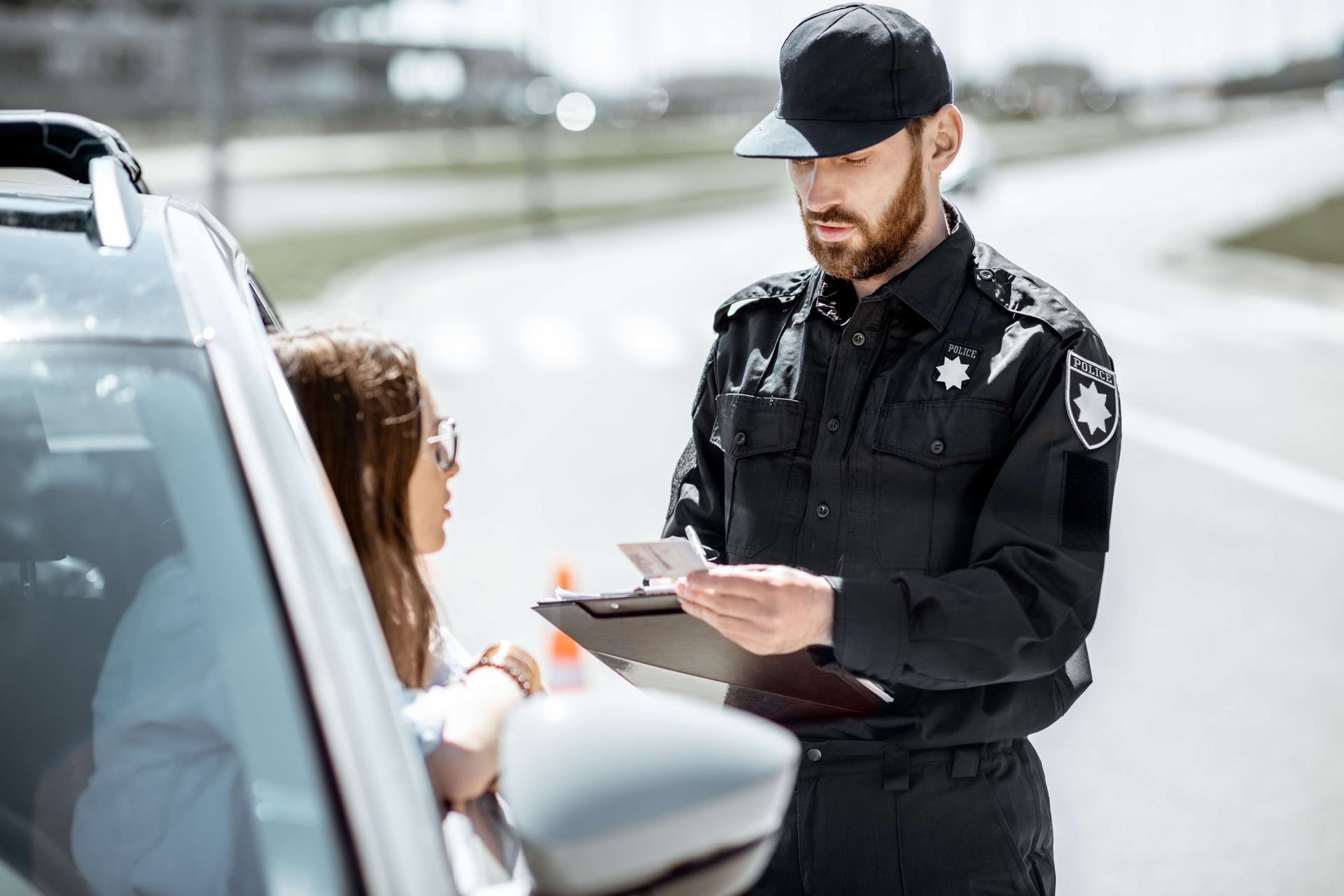 Police officer writing a ticket beside a car while speaking with a driver
