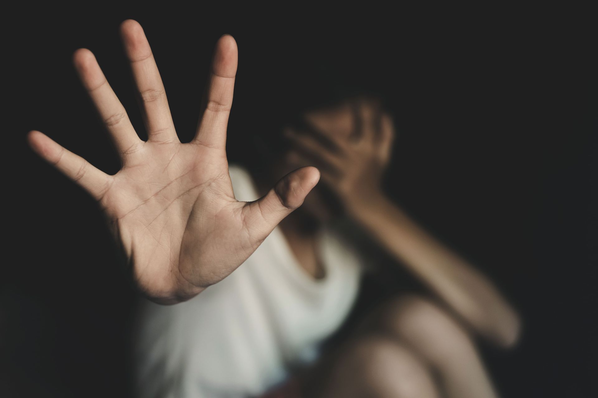 Person with raised hand in foreground, another blurred figure sitting behind in a dark room