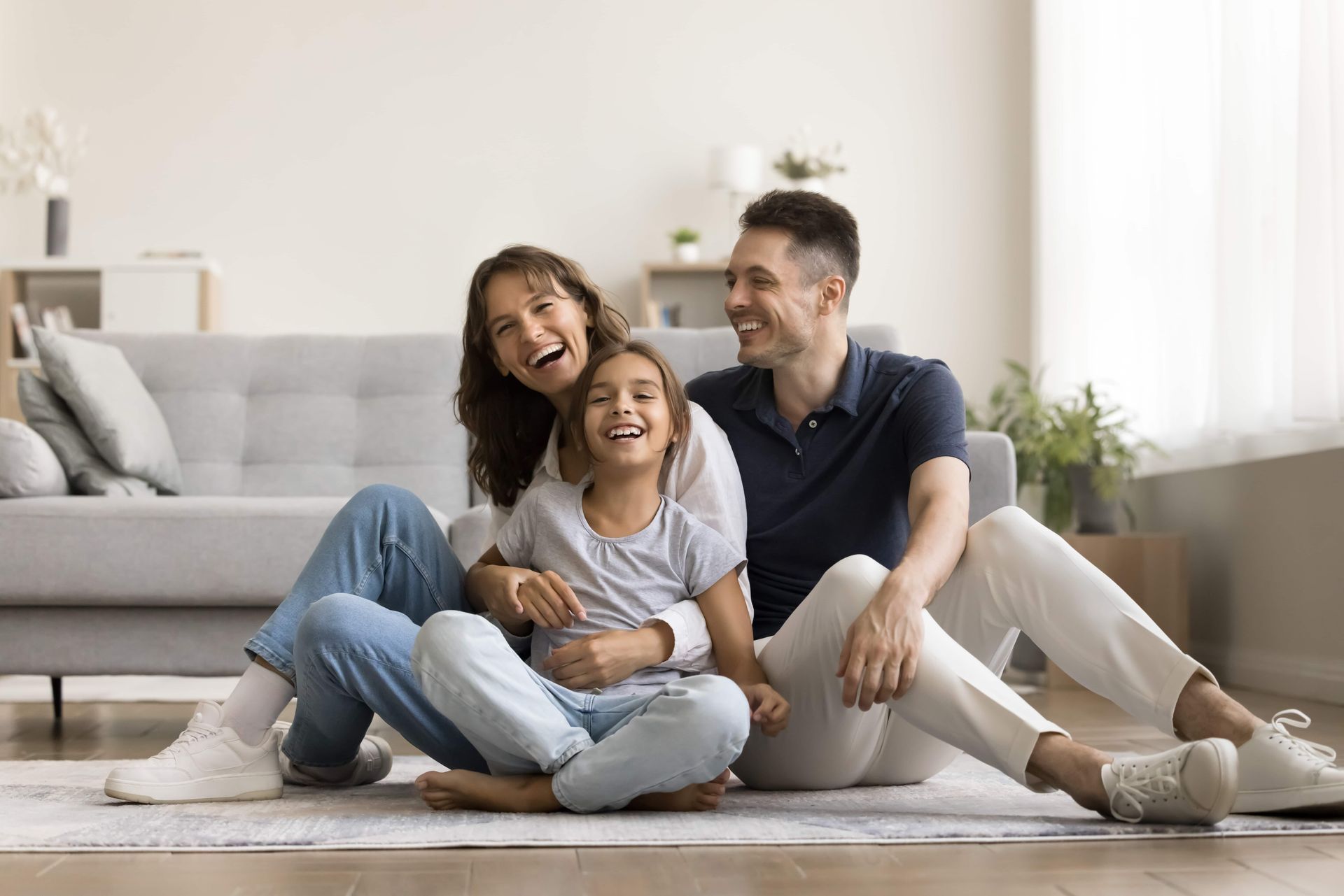 Three people sitting on a living room floor, smiling and leaning together by a gray sofa.