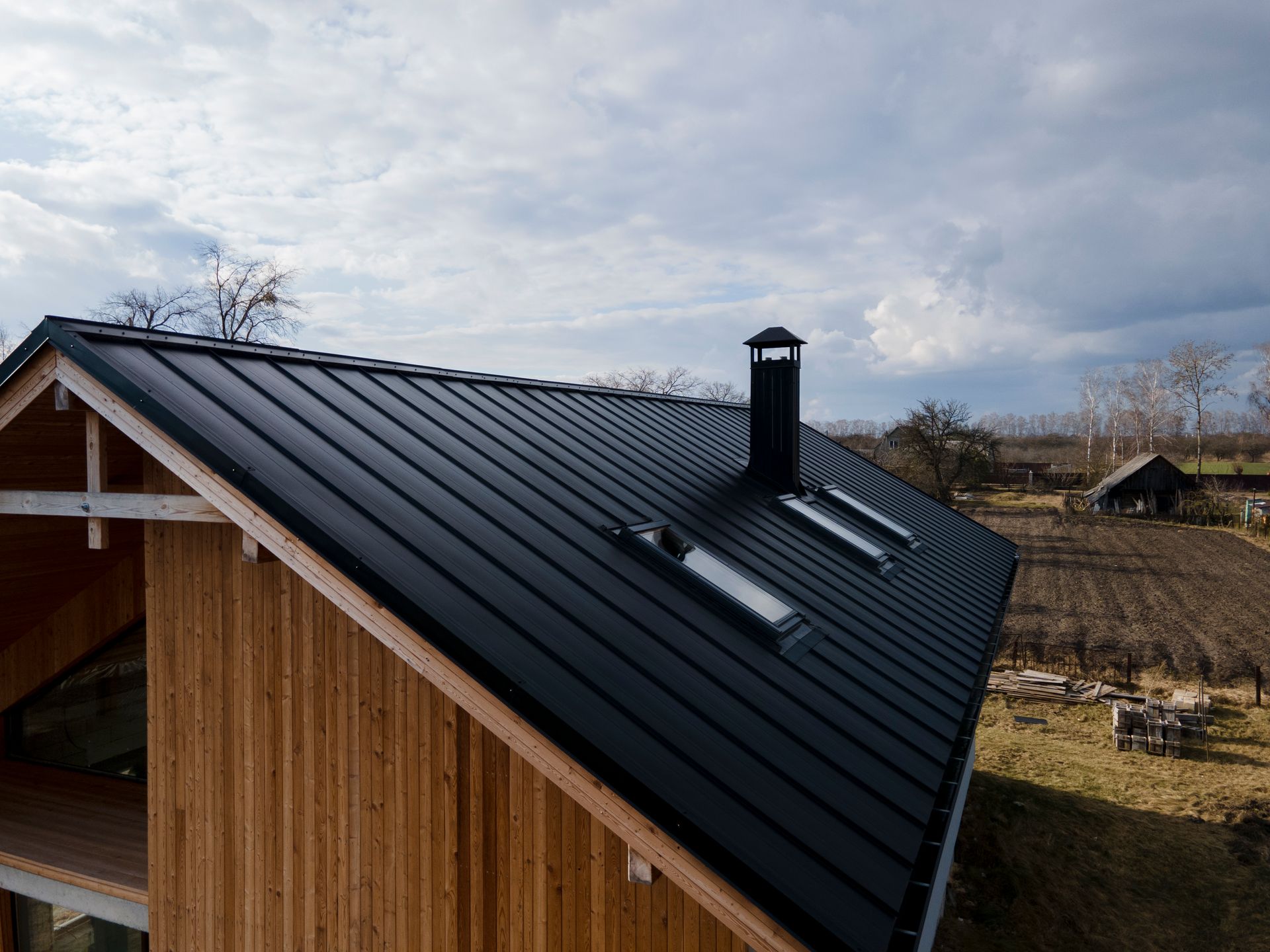 Black metal roof on a wooden house with three skylights, chimney, and a small barn in the background.