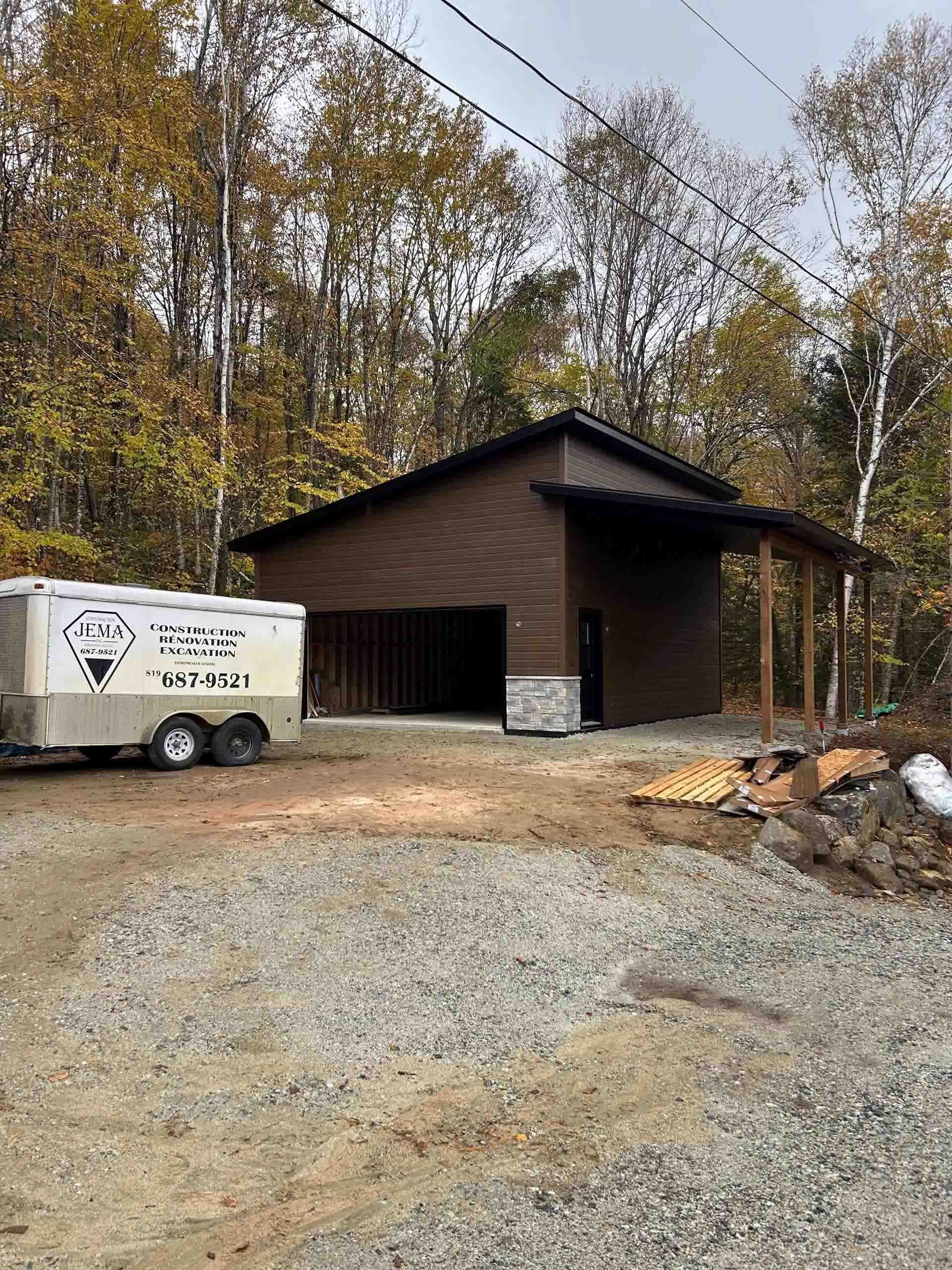 Un hangar brun avec une base en pierre et un garage ouvert se trouve sur un terrain gravillonné à côté d'une remorque cargo blanche dans une zone boisée.