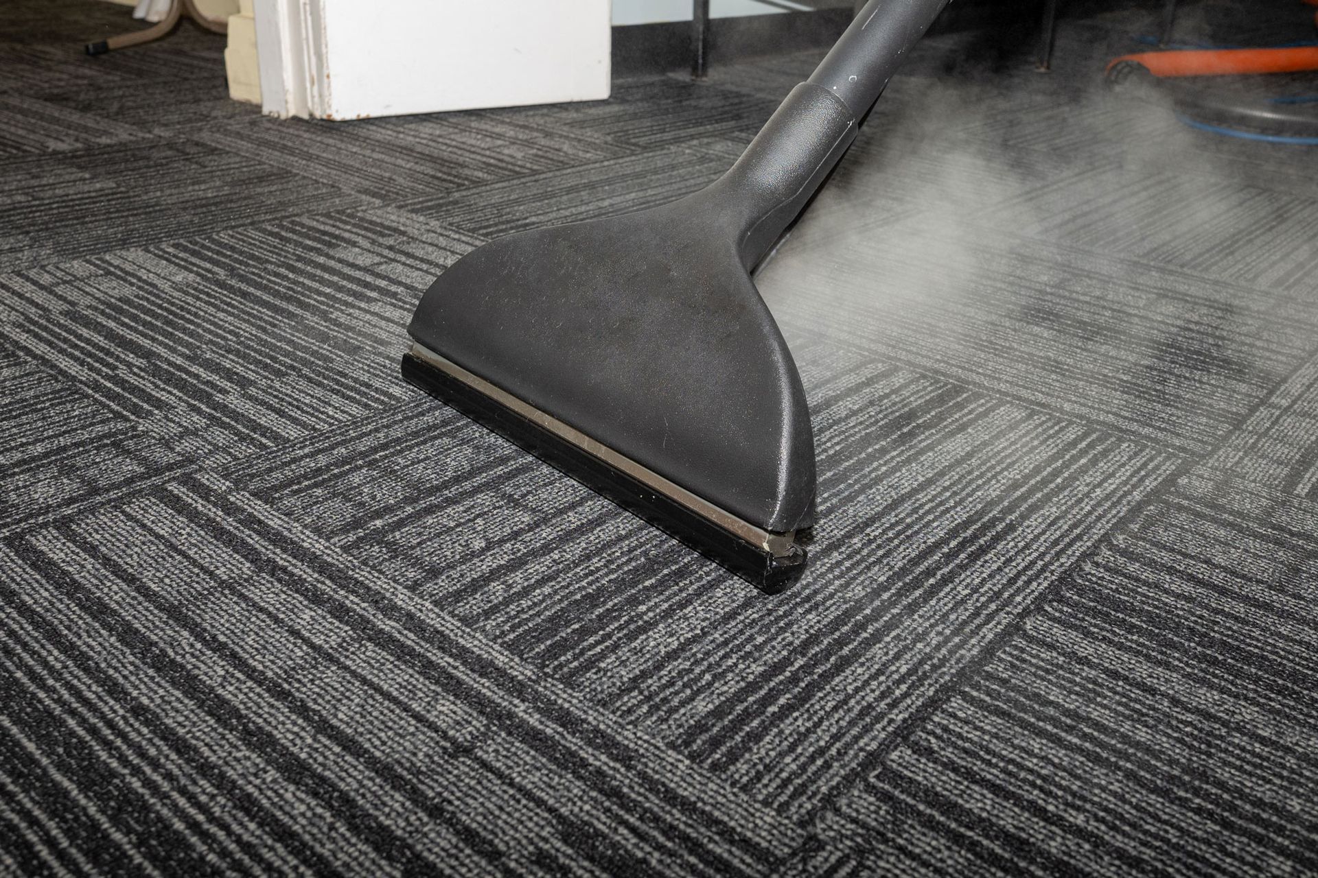 Carpet being steam cleaned with a black nozzle, releasing steam over dark patterned carpet.