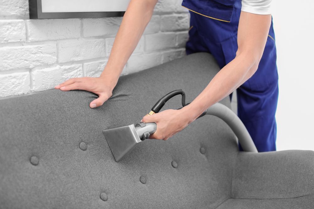 Person in blue uniform cleaning a grey sofa with a vacuum in a room with a white brick wall.