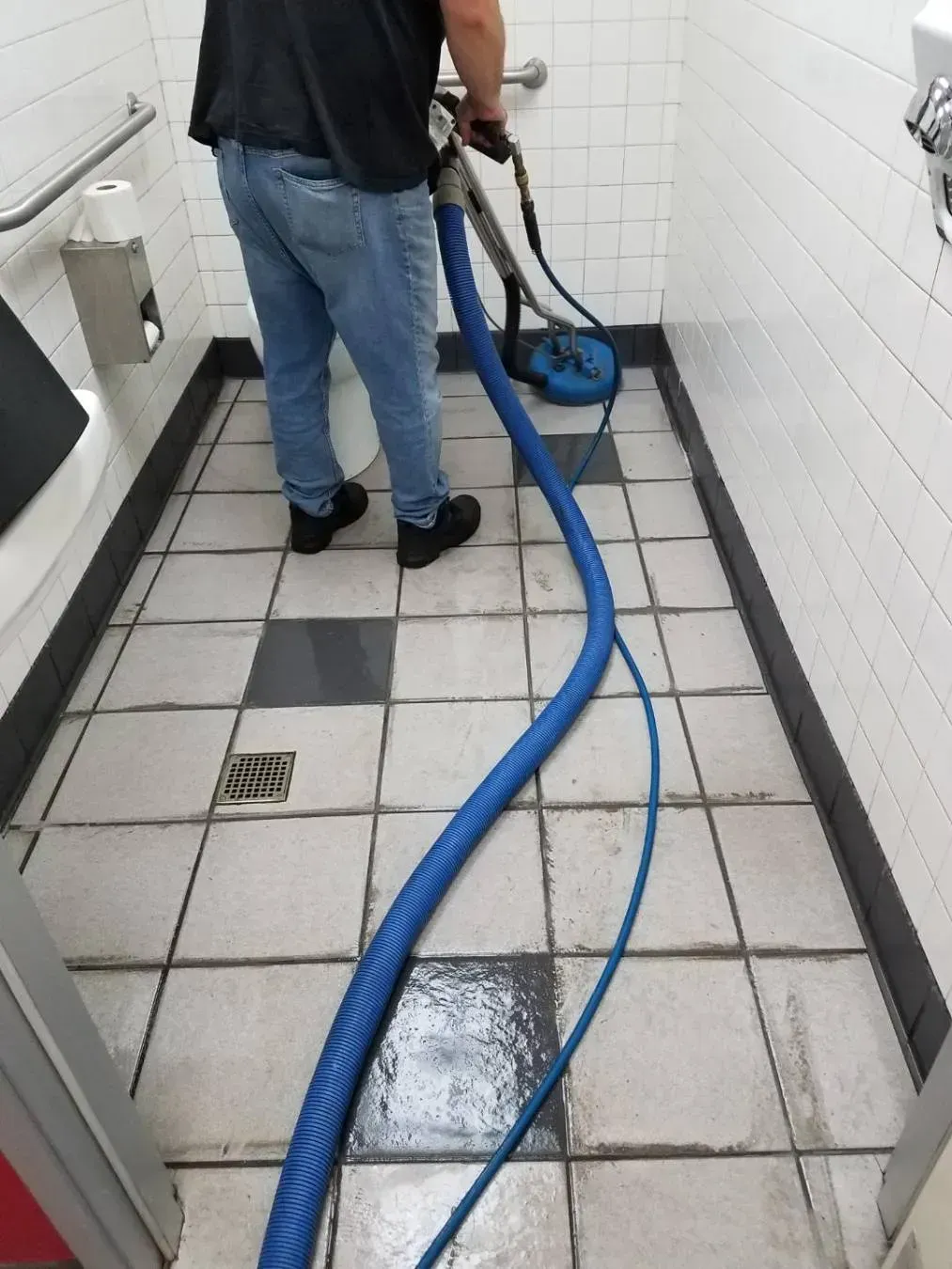 Person cleaning bathroom floor with a blue hose and cleaning tool. White tiled walls and floor.