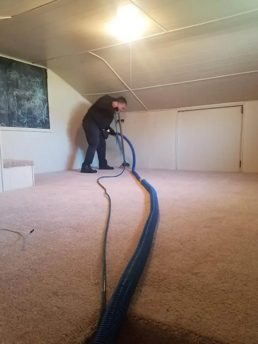 Man cleaning a tan carpeted room with a blue hose; room has white walls and a single overhead light.