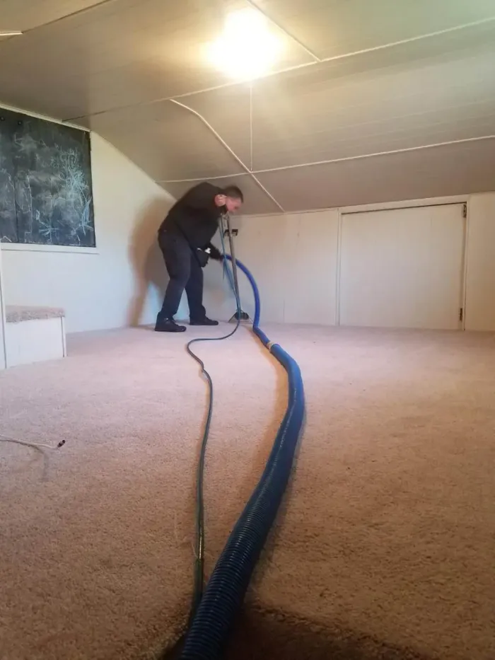 Man cleaning a tan carpeted room with a blue hose; room has white walls and a single overhead light.