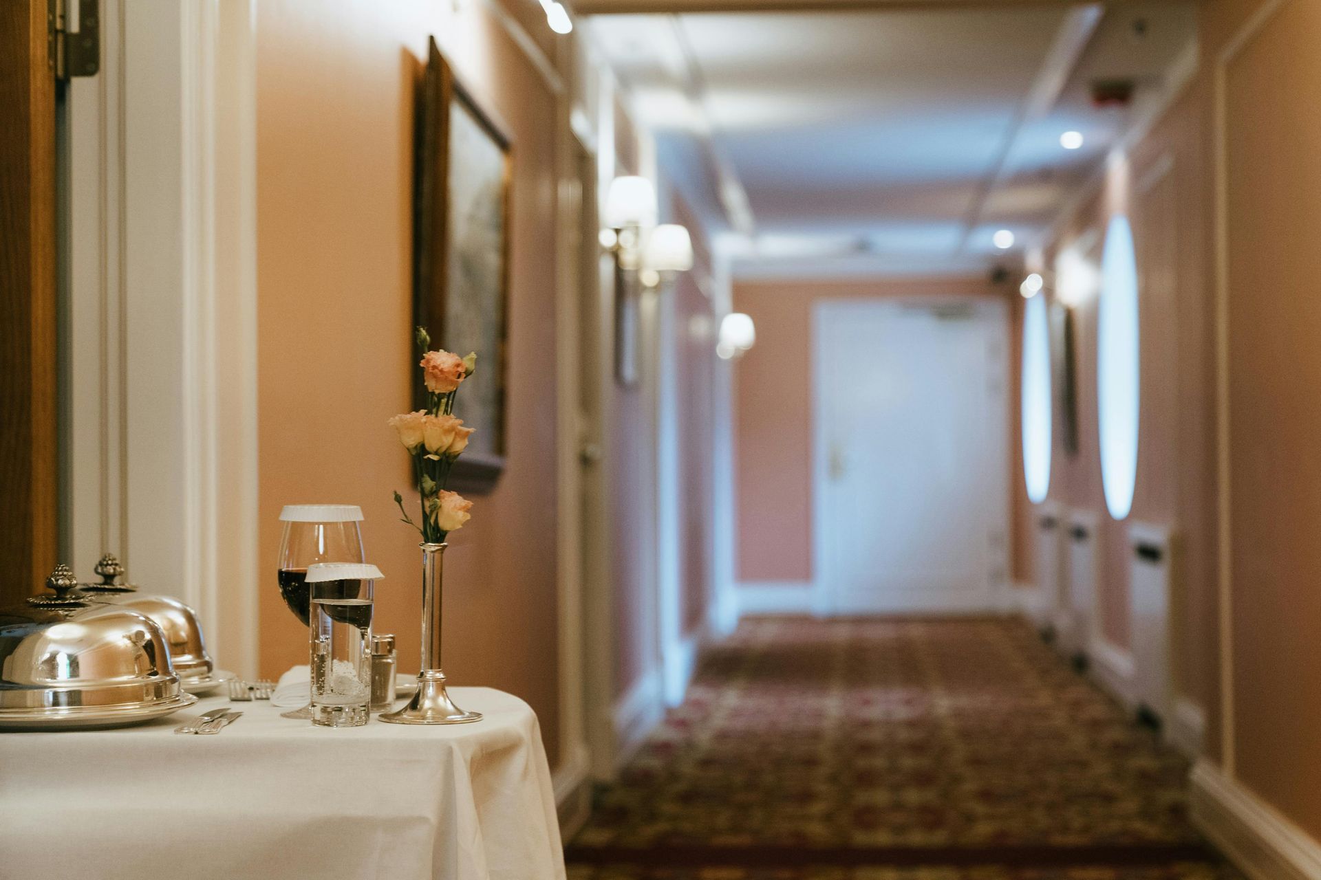 Hotel hallway with table set with glassware, flowers and serving dishes; pale peach walls, patterned carpet.
