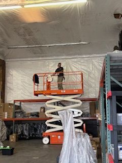 A person stands in an orange scissor lift, working on a warehouse ceiling. The warehouse has metal shelving.
