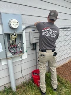 An electrician in a gray shirt works on wiring outside a building. He is wearing a cap and light tan pants.