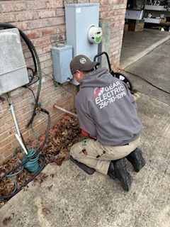 Electrician kneels to work on an electrical box on a brick building. He wears a gray hoodie with logo and khaki pants.