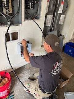 Electrician working on electrical panel, wiring a new box on a white wall. Gray shirt, baseball cap, and tools visible.