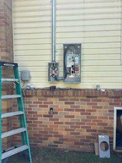 Exterior electrical work in progress: a metal electrical box on a brick and siding wall, ladder nearby.