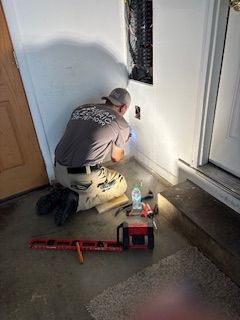 An electrician kneels near an electrical panel, working with tools in a corner near a door.