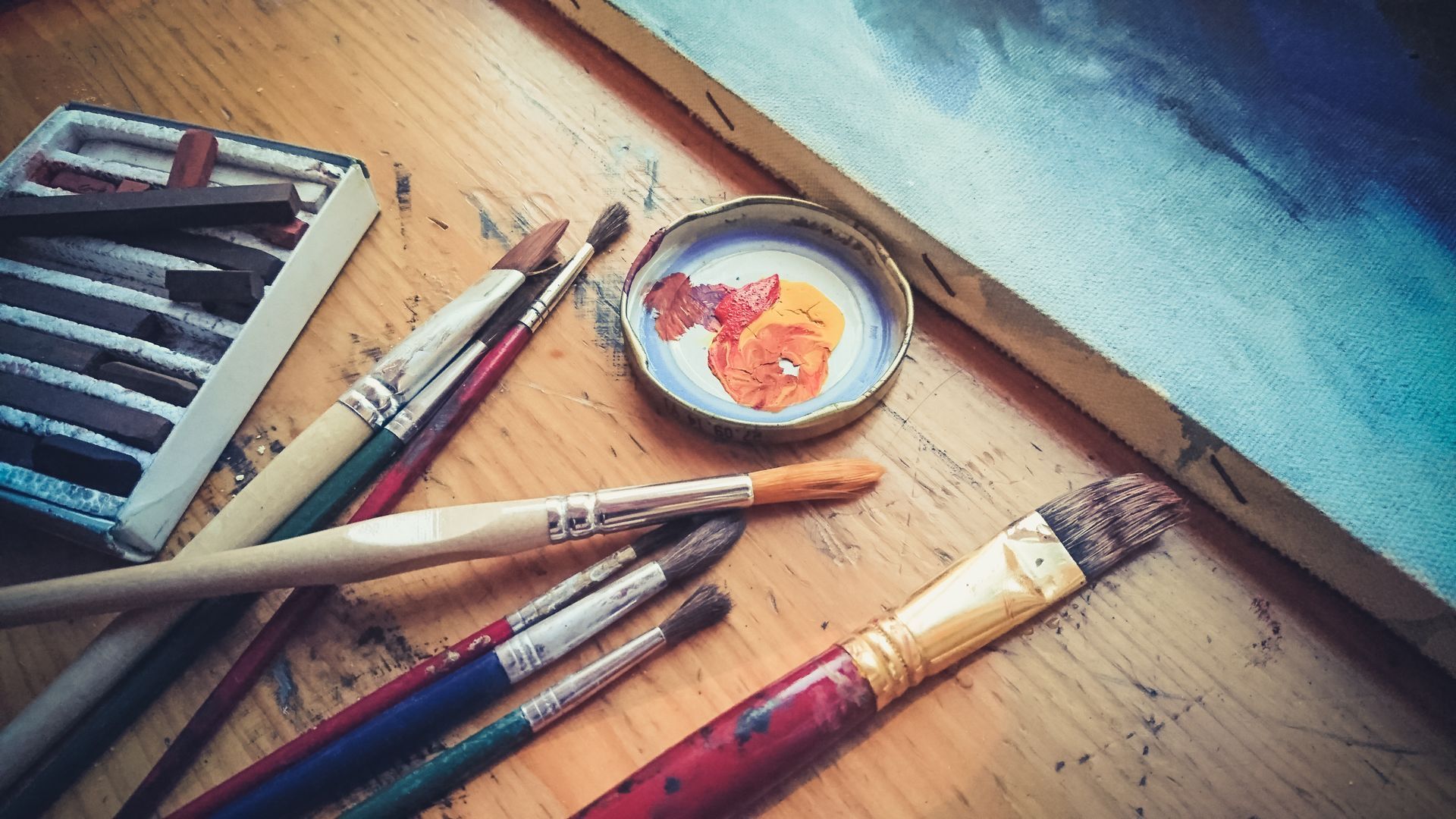 Pinsel, Pastellkreiden und ein mit Farbe gefüllter Deckel auf einer Holzfläche neben einer Leinwand