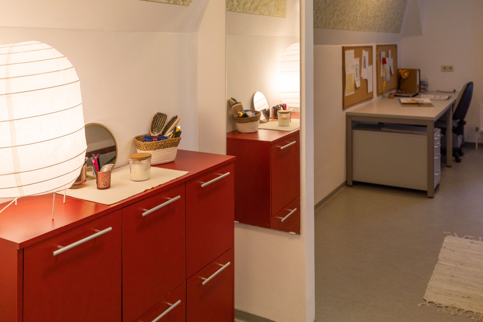 Im Vordergrund ein roter Schrank mit weißer Lampe, Spiegel und Toilettenartikeln, daneben  Schreibtisch und Stuhl.
