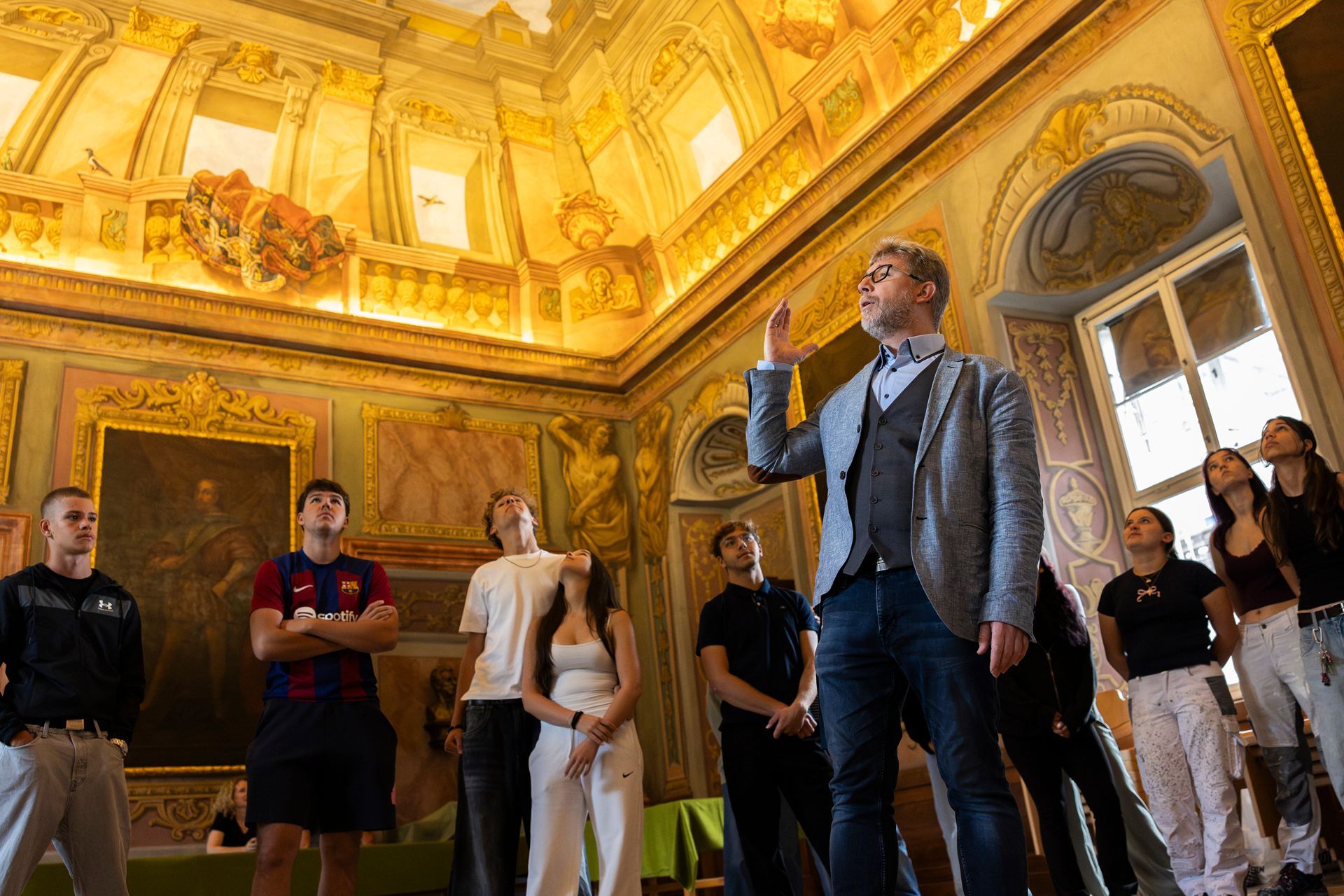 Man gestikuliert, während eine Gruppe in einem vergoldeten Raum zu einer bemalten Decke hinaufblickt.