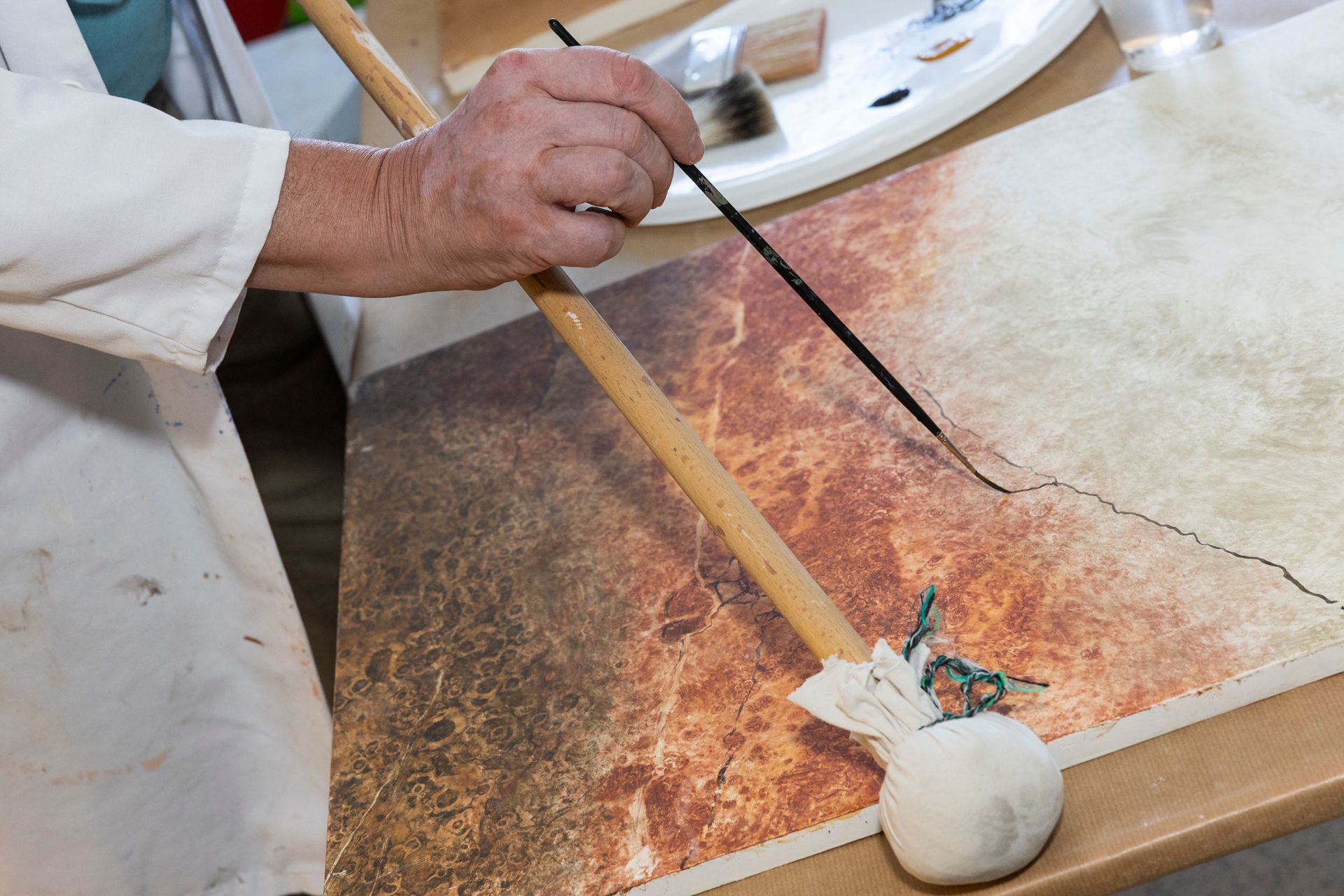 Eine Person malt mit Pinsel auf Papier; Holzgriff und Farbpalette sind sichtbar.