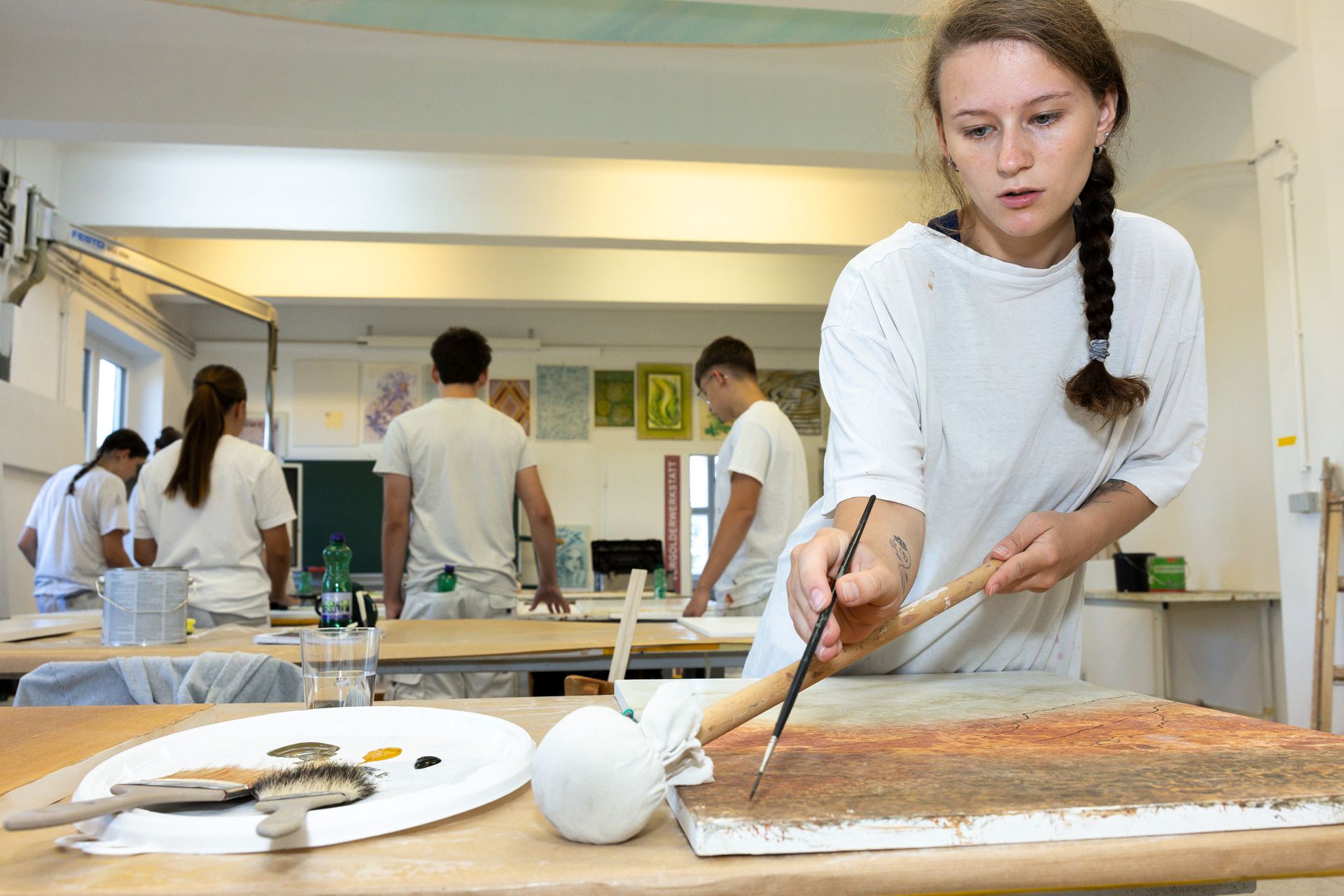 Eine Frau malt mit Pinsel an einem Tisch, Atelierkulisse. Im Hintergrund sind weitere Studenten zu sehen.