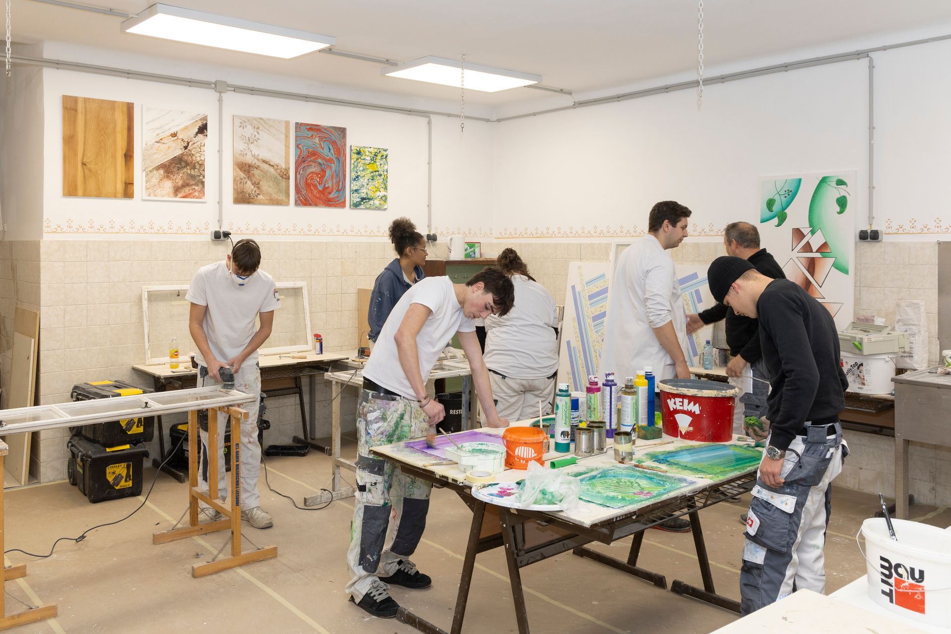 Studenten in Arbeitskleidung üben Maltechniken an Tischen in einem hell erleuchteten Atelier, an  Wänden Kunstwerke hängen.