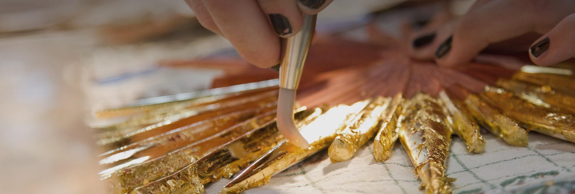 Nahaufnahme von Händen, die mit Blattgold arbeiten und es mit einem Werkzeug auf eine Oberfläche auftragen.