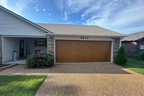 A single-story suburban house with a brown garage door, light-colored siding, and a gravel driveway under a sunny sky.