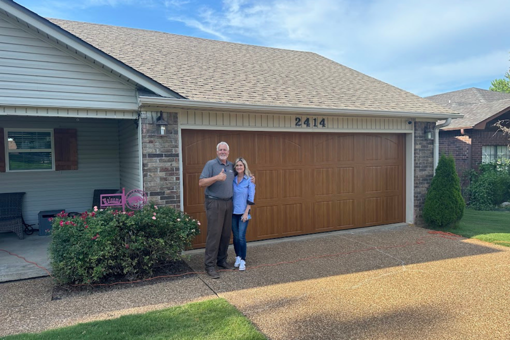 A man and a woman stand together in front of a house with a brown garage door and a gravel driveway.
