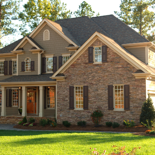 Two-story house with green siding, brown stone, and dark brown shutters, set against a green lawn and trees.