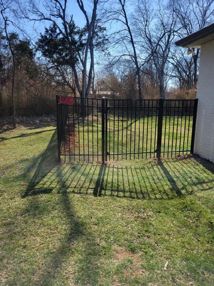 A black fence is surrounding a lush green yard with trees in the background.