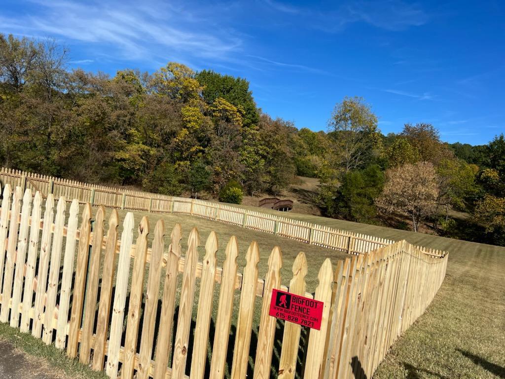 A wooden picket fence surrounds a field with trees in the background.