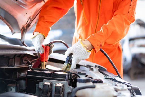 Mechanic in orange jacket using jumper cables on a car battery under the hood
