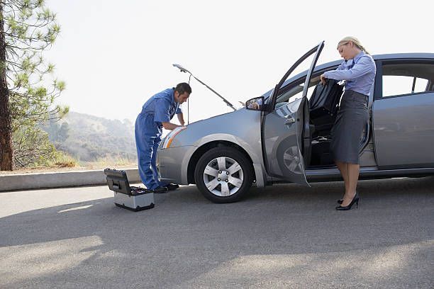 Two people inspecting a gray car with its hood open and a tool box on the road by a hillside.