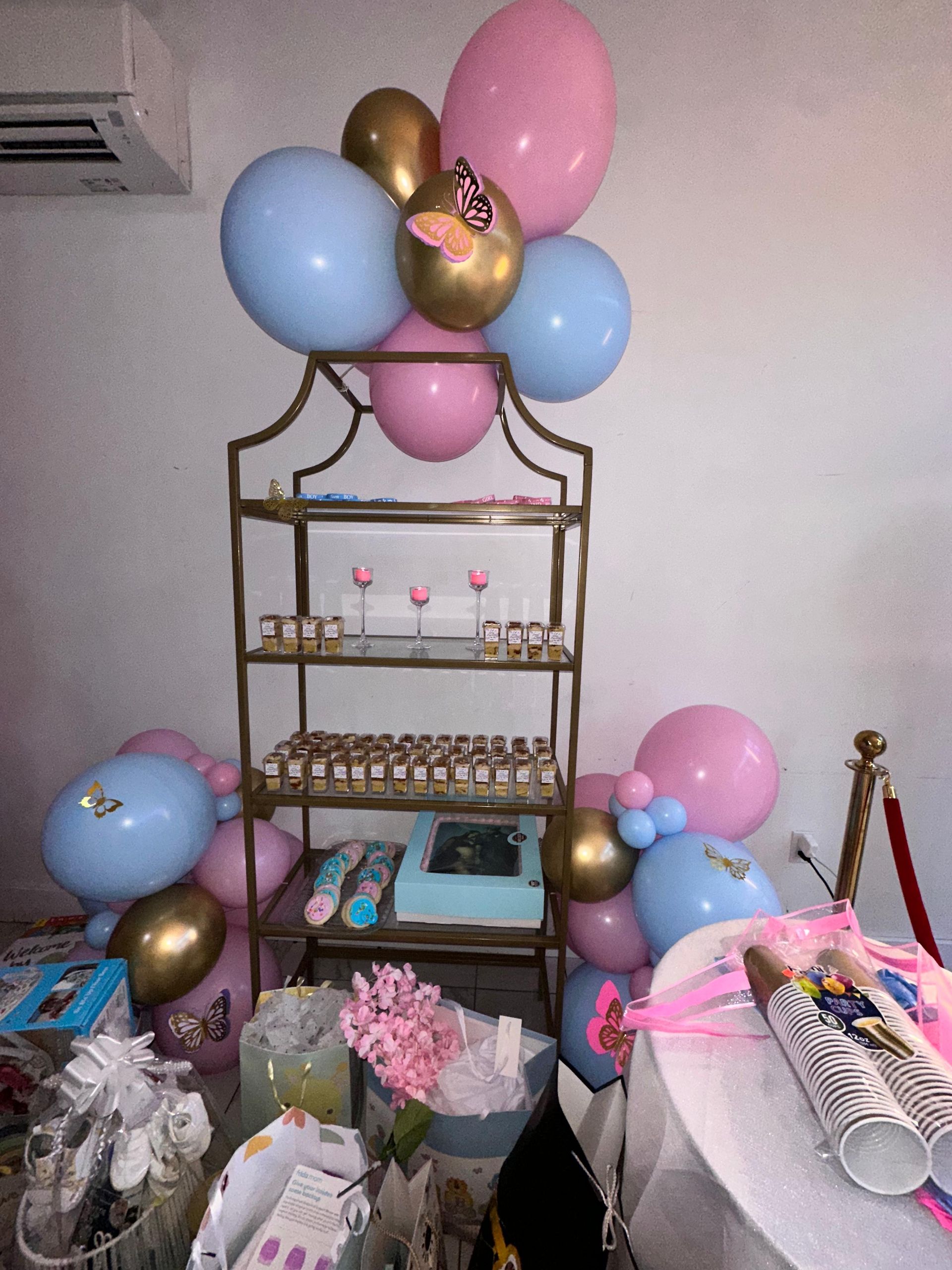 A gold tiered display stand decorated with blue, pink, and gold balloons, holding party favors and treats.
