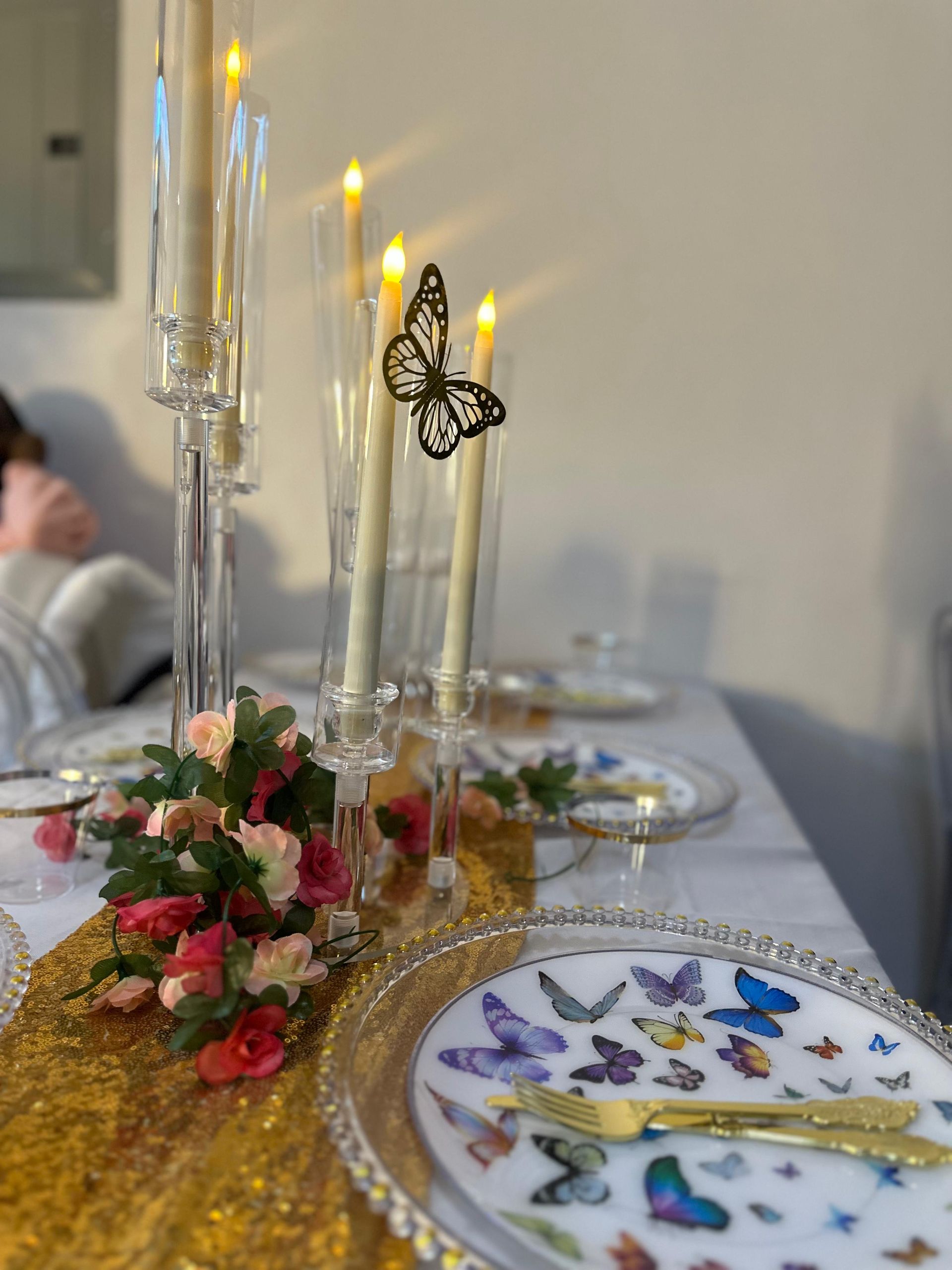 A table set with a gold runner, butterfly-patterned plates, candles in glass holders, and a small pink floral arrangement.