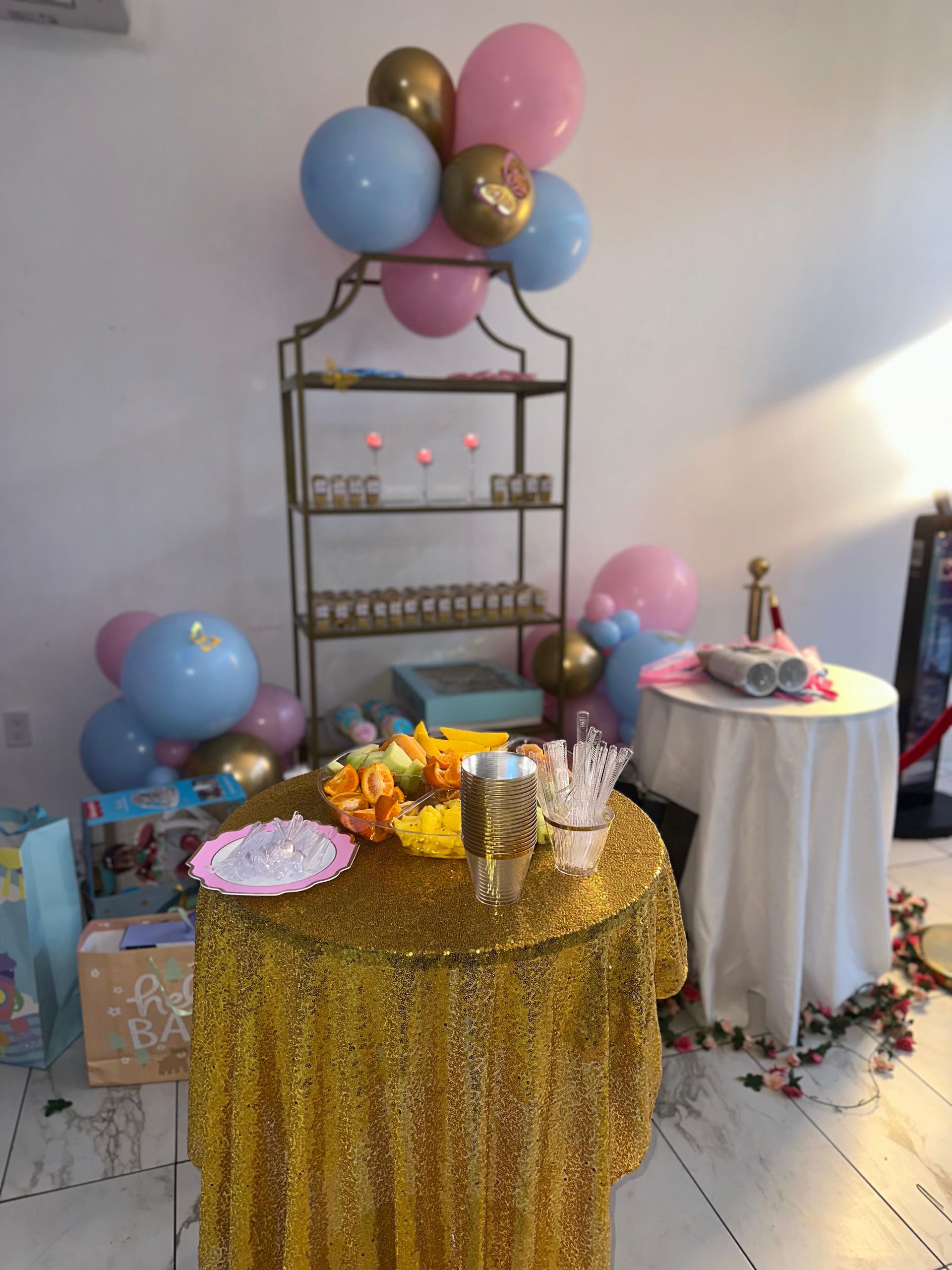 A display of balloon clusters in pink, blue, and gold above a gold sequin-covered table and a tall metal shelf.