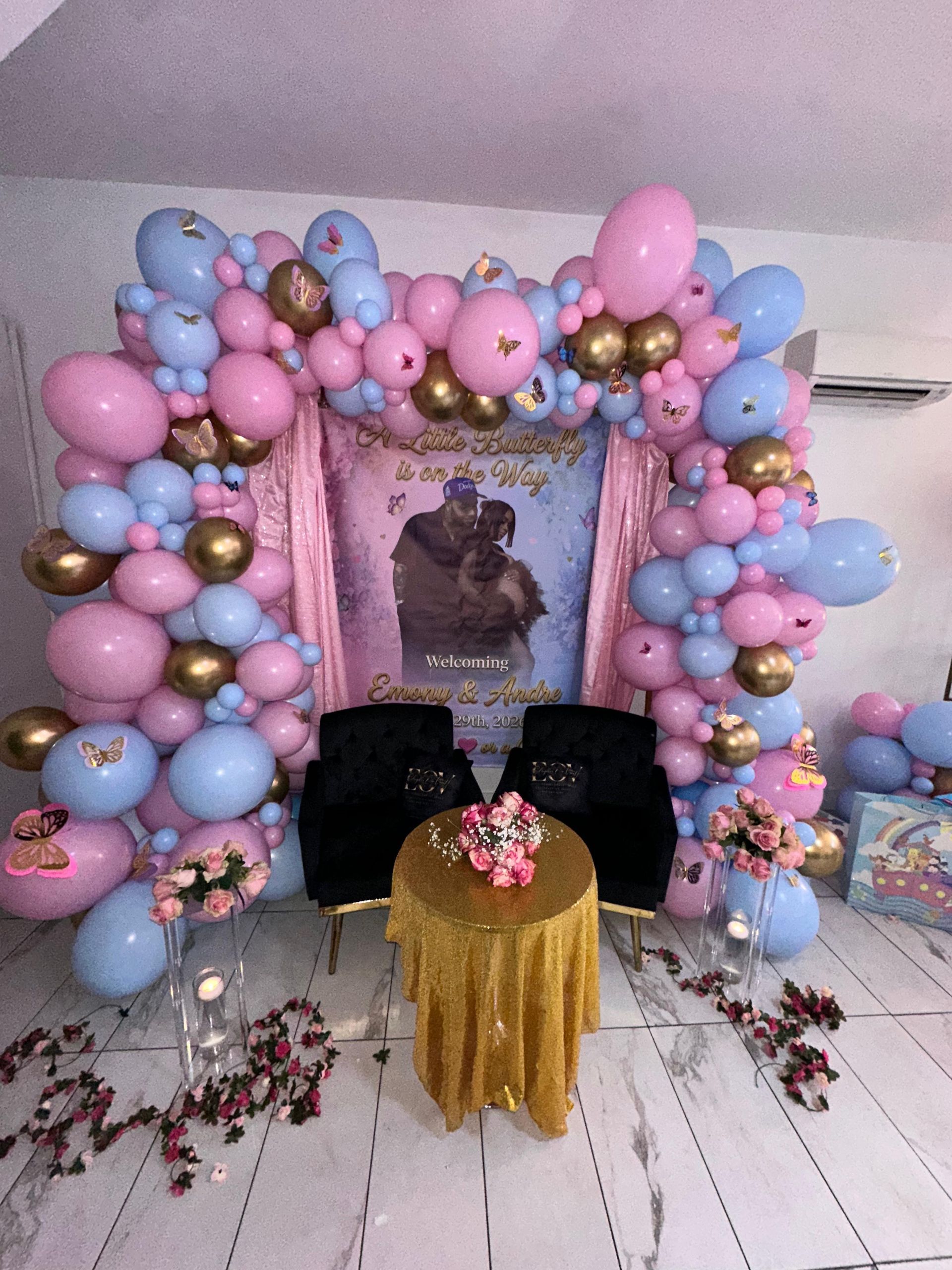 A party backdrop with a balloon arch in pink, blue, and gold surrounding a central poster, two black chairs, and a table.