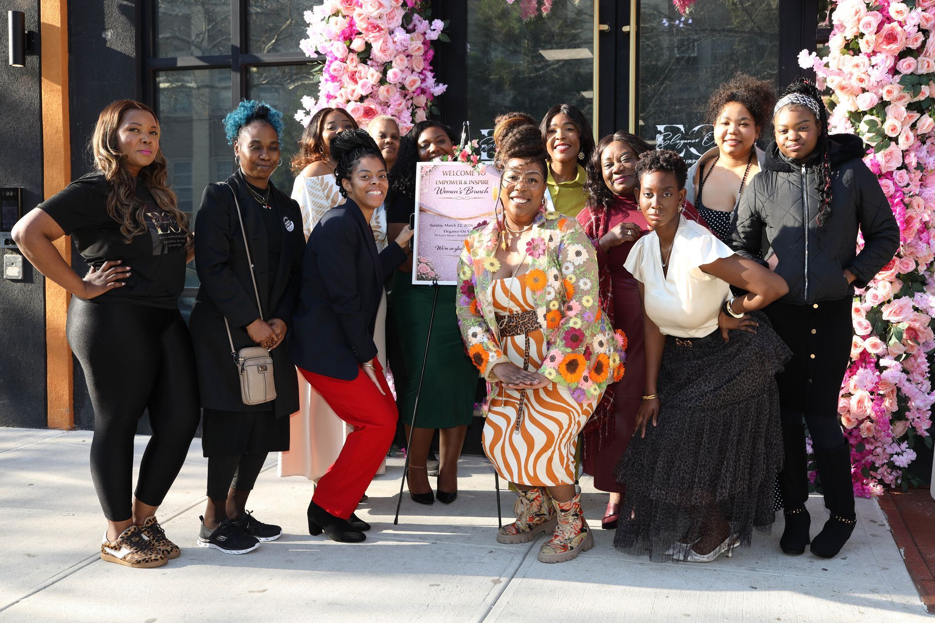 A group of eleven people stand posing together outside, holding a sign in front of a building decorated with floral arches.