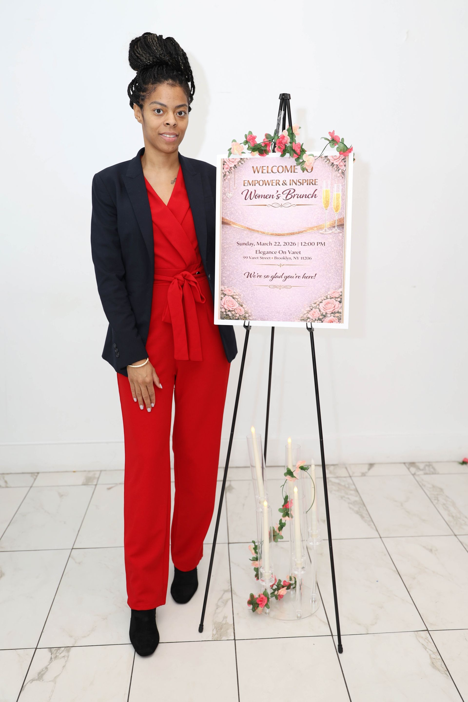 A person wearing a dark blazer and red jumpsuit stands next to a floral-decorated sign on an easel in a white room.