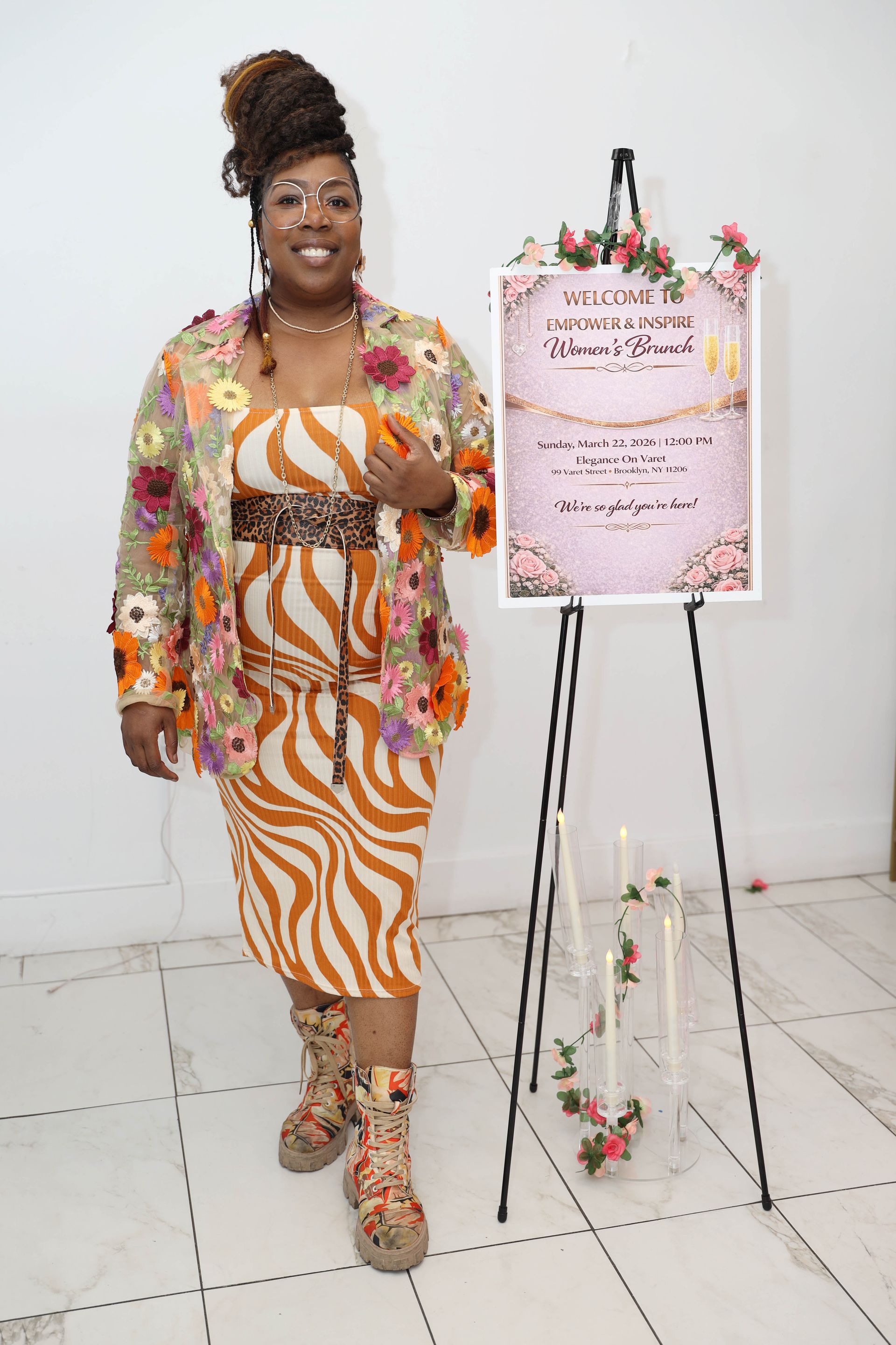 A person smiling next to an easel displaying a floral-themed event sign, wearing a patterned dress, jacket, and boots.