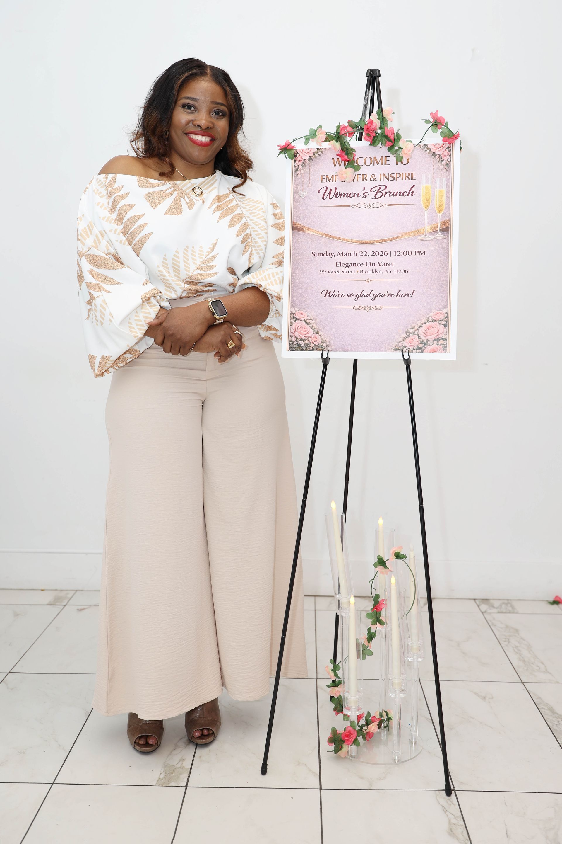 A smiling person stands next to a floral-decorated sign on an easel in a bright, white-walled room.