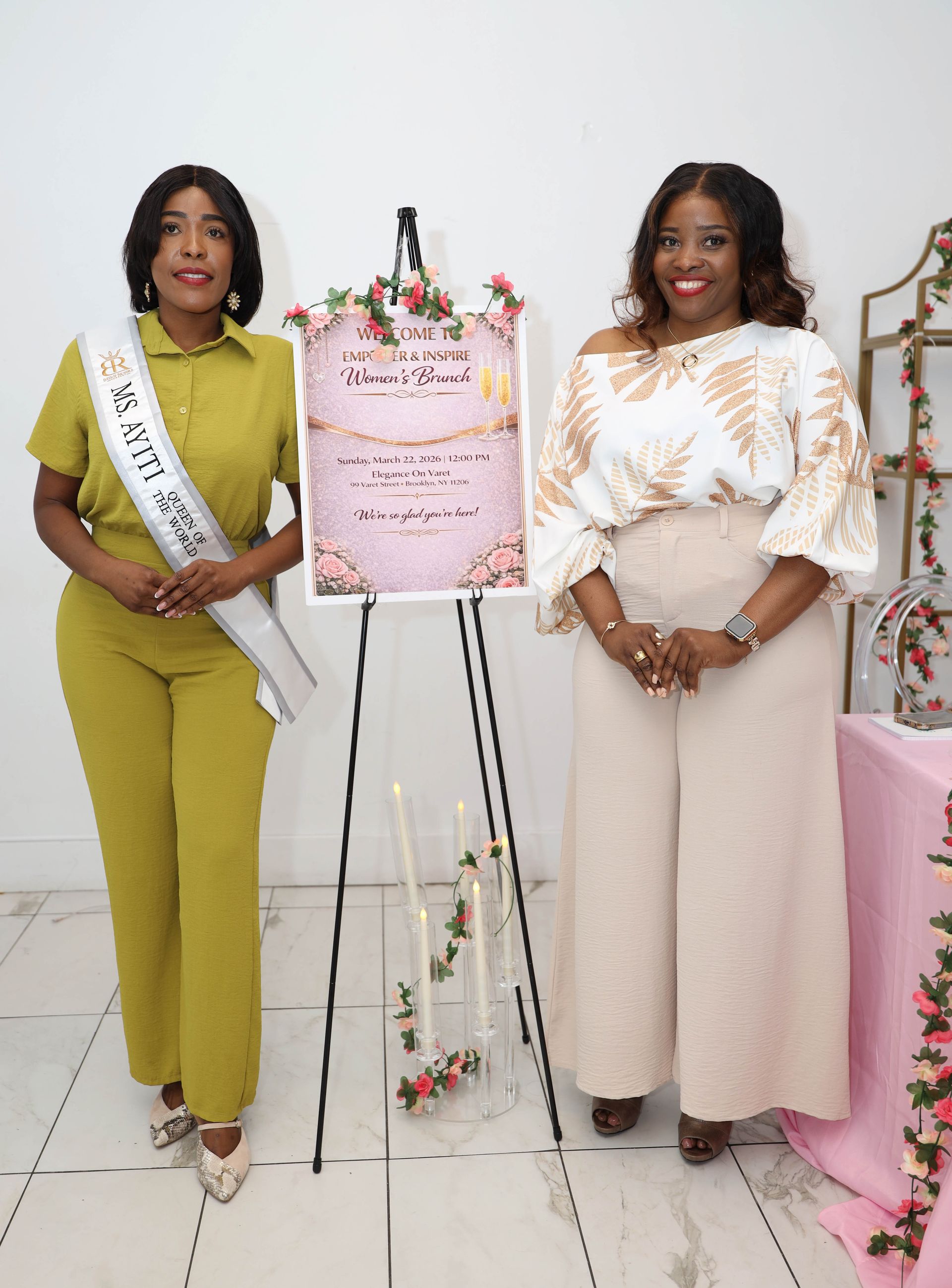 Two people stand on either side of a decorative sign on an easel in a bright room with pink accents.